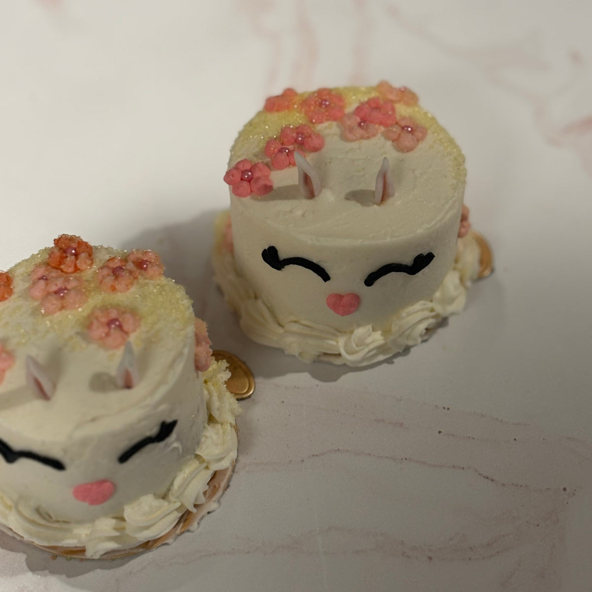 Two small bunny cakes with facial features and floral decorations on a marble surface.
