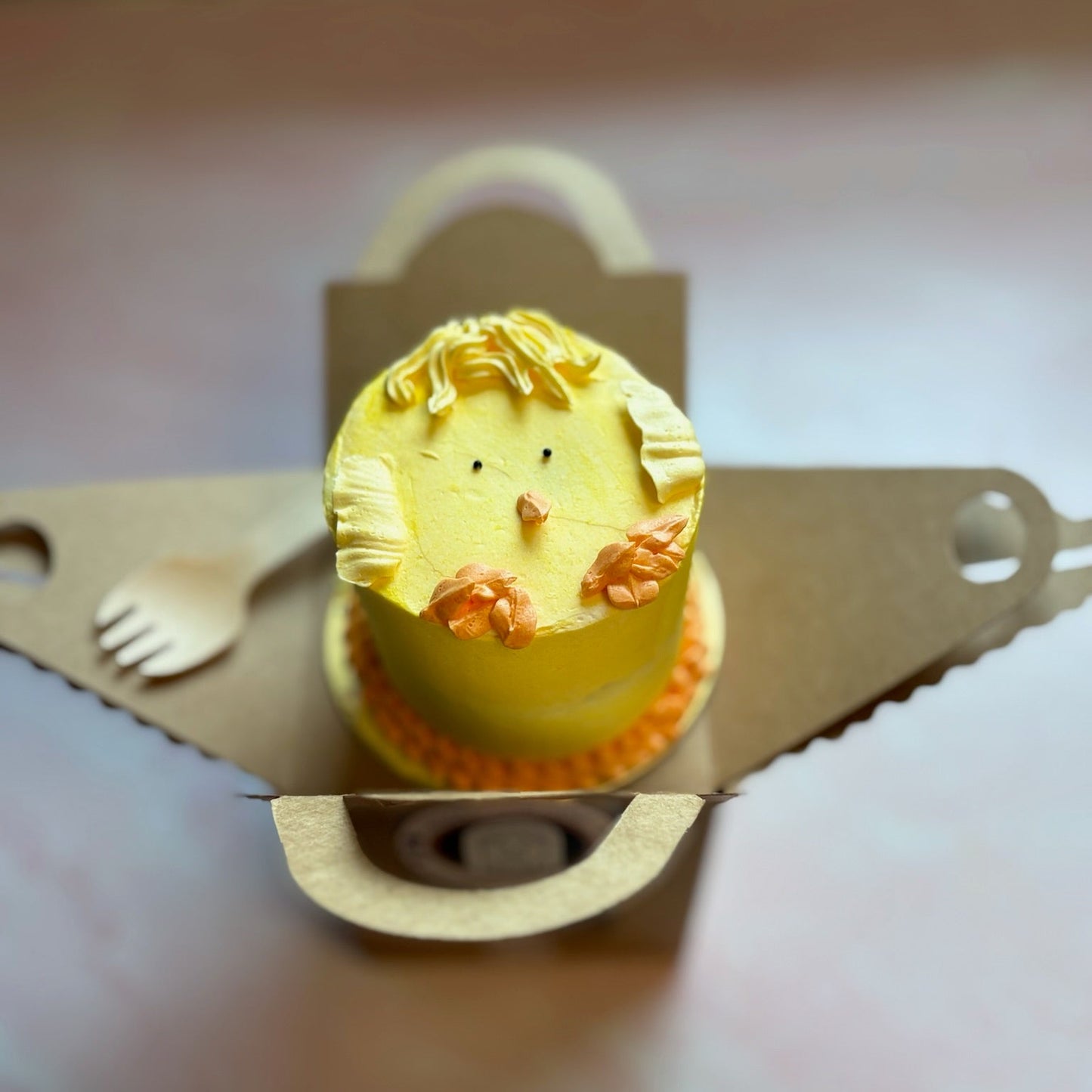 Small yellow cake with a baby chick design on top, placed on a cardboard mini cake box