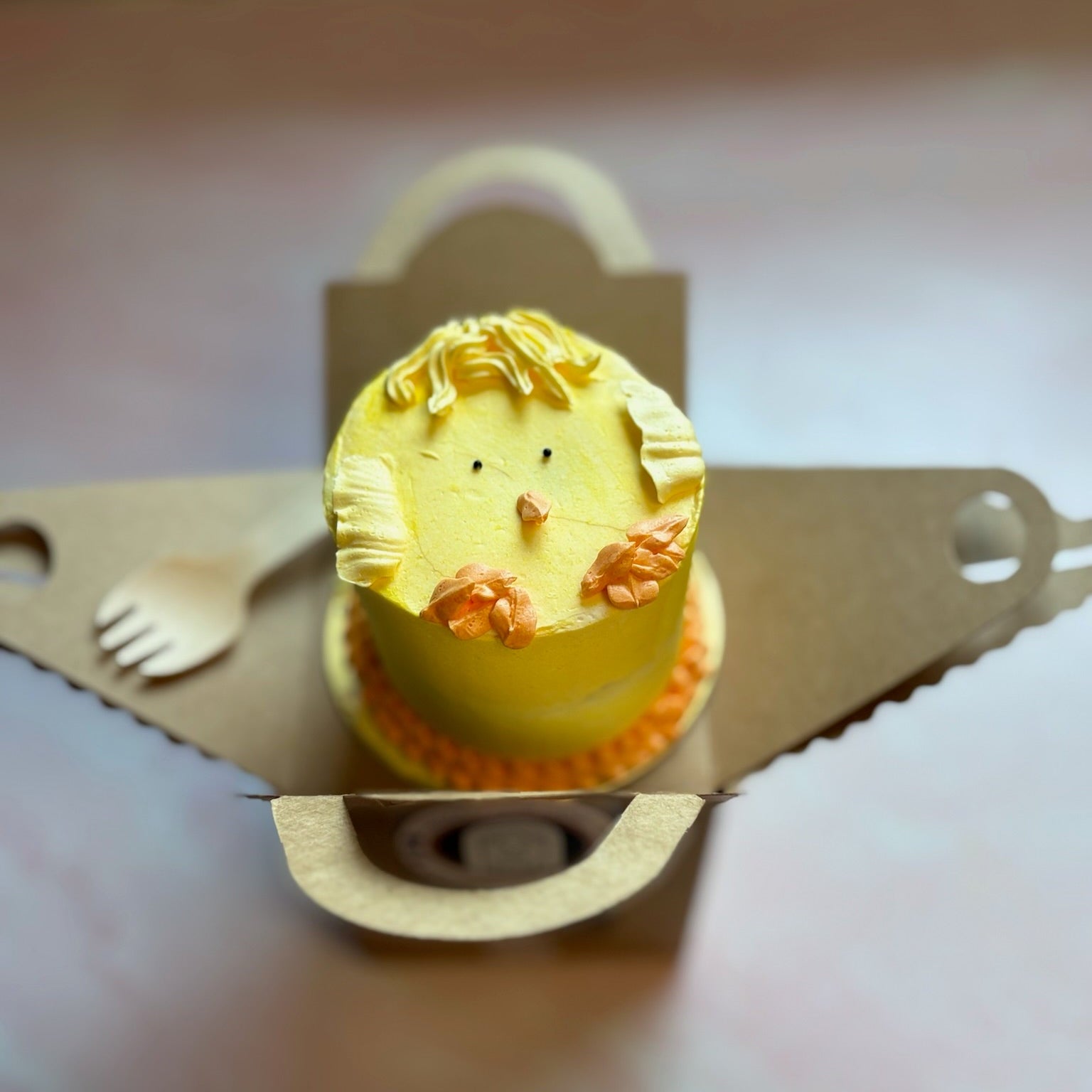 Small yellow cake with a baby chick design on top, placed on a cardboard mini cake box