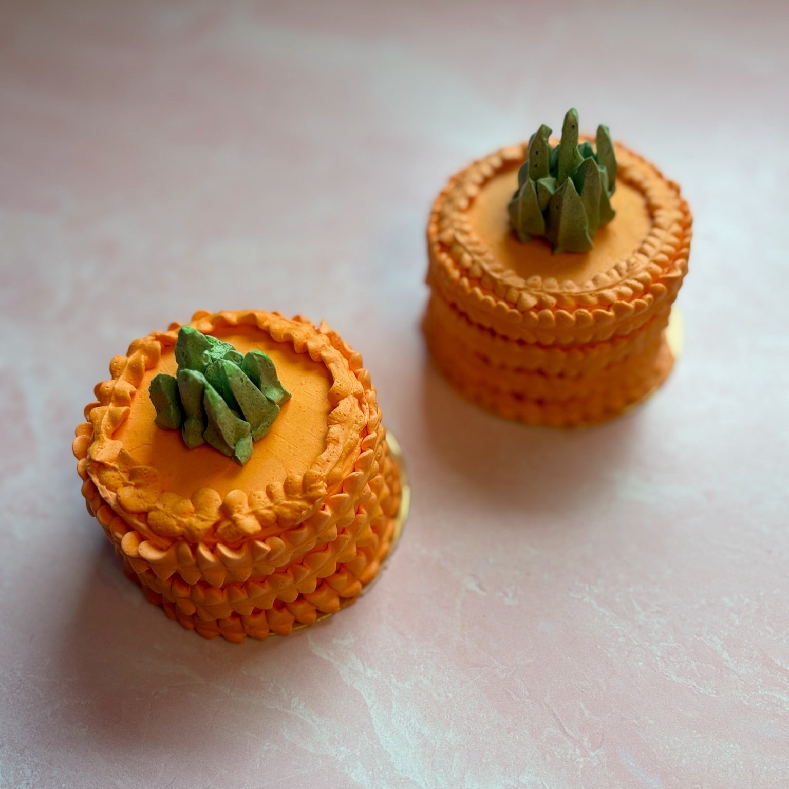 Two small orange cakes with green decorations, piped to resemble a carrot, on a light pink surface